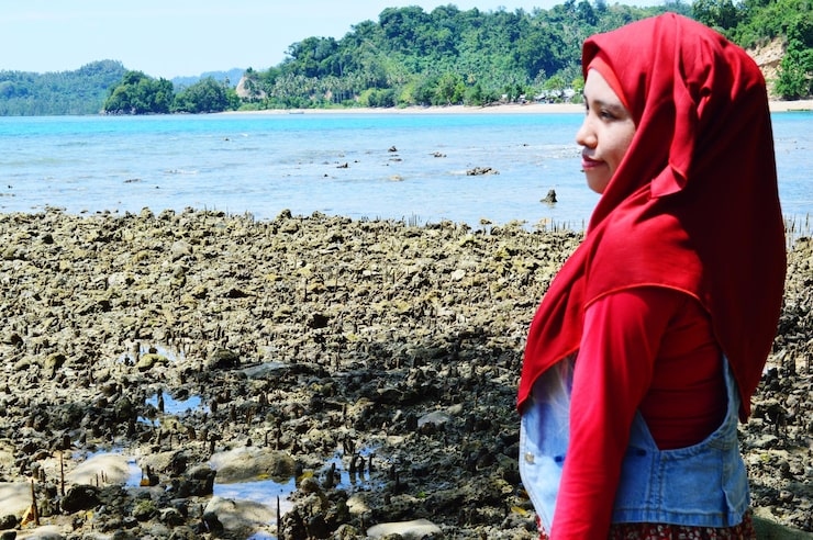1 A woman in a red hijab and red top with a denim vest, standing on a rocky shore by the sea.jpg