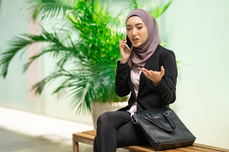 businesswoman in hijab talking on phone sitting on bench with bag and plants in background.png