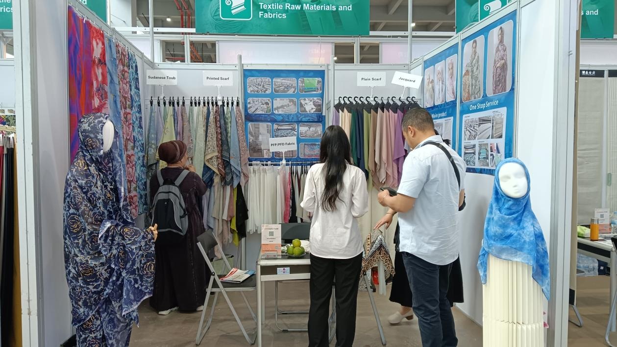 a textile exhibition booth displaying various scarves and fabrics with people browsing.jpg