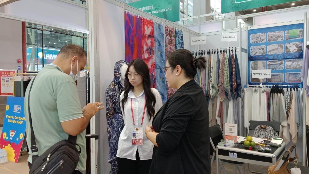people at a textile exhibition booth discussing with various printed scarves and fabrics on display.jpg